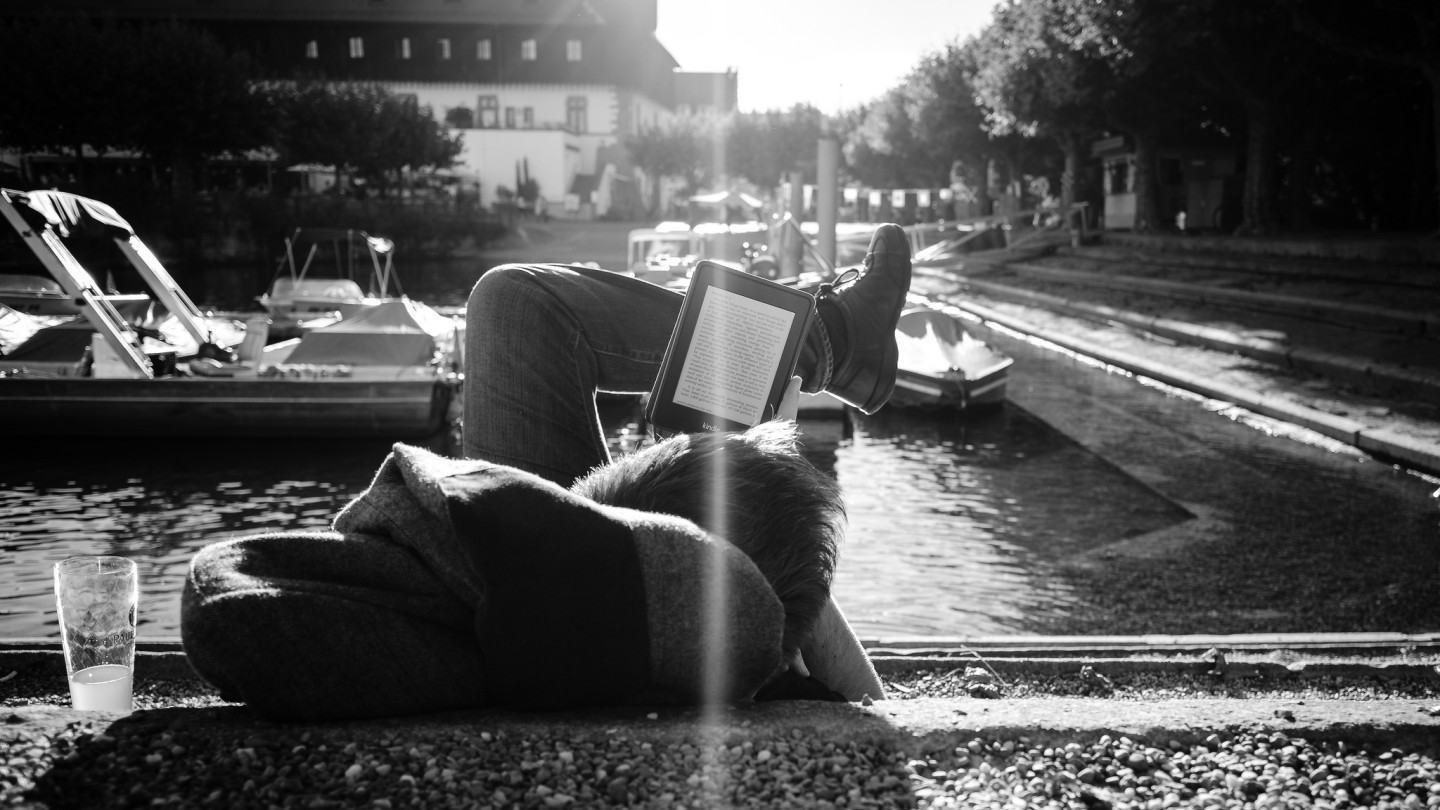 Pessoa lendo um Kindle deitada na beira do lago ou rio.