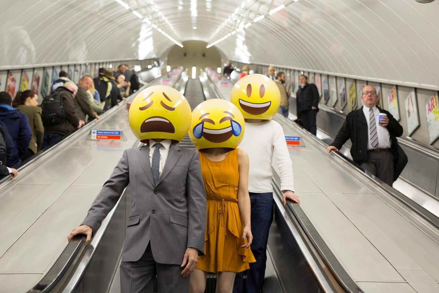 Três pessoas com cabeças de emoji descendo escadas no metrô de Londres.
