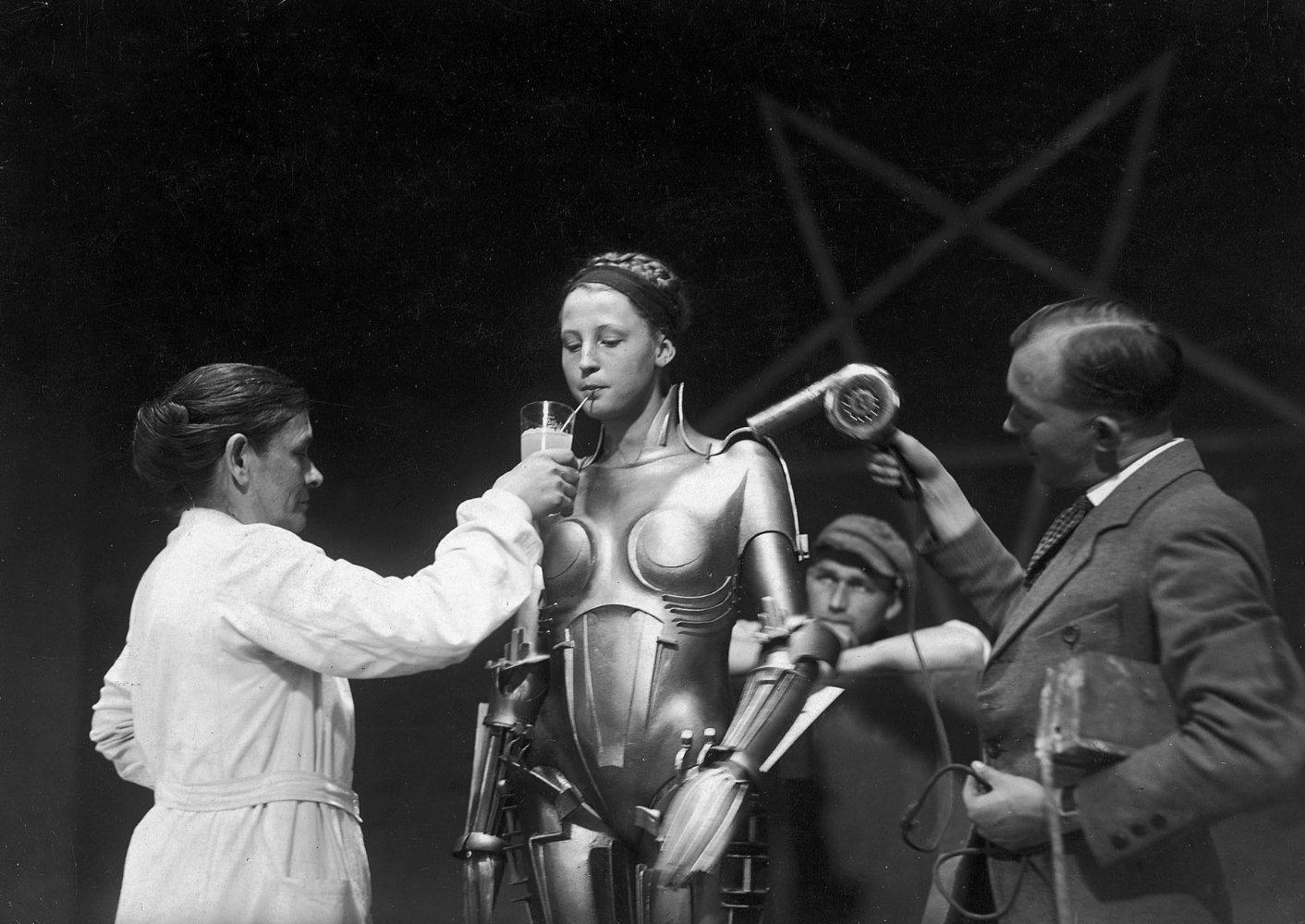 Brigitte Helm na pele da robô, sem o capacete, sendo auxiliada por duas pessoas para se alimentar e se refrescar dentro da roupa/armadura.