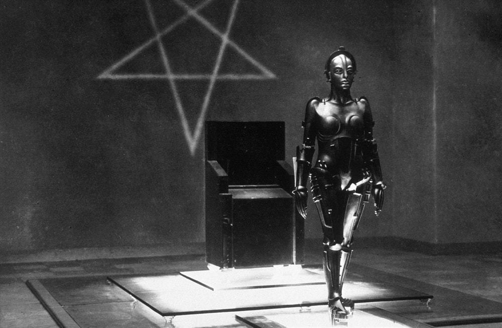 Foto em preto e branco do robô do filme “Metrópolis” de pé, andando. Ao fundo, uma poltrona e uma estrela de cinco pontas desenhada na parede.