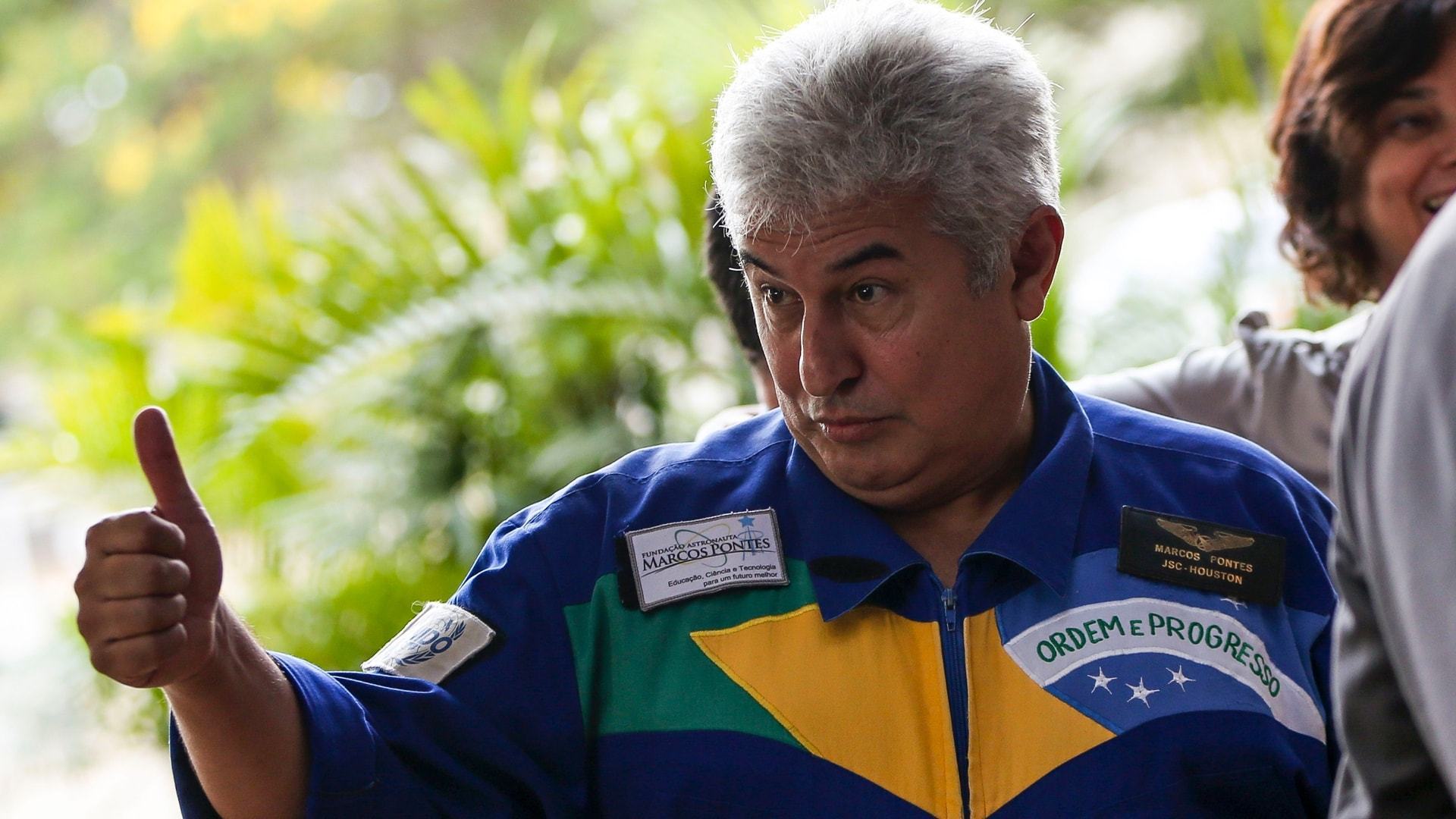 Ministro Marcos Pontes, em seu famoso traje nas cores da bandeira do Brasil, fazendo um sinal de positivo.