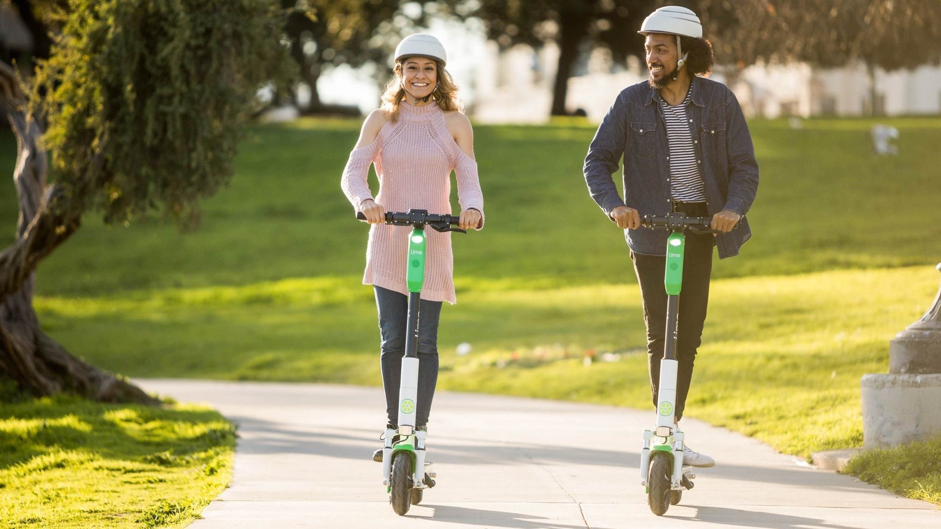 Duas pessoas andando em patinetes elétricos em um parque no amanhecer.