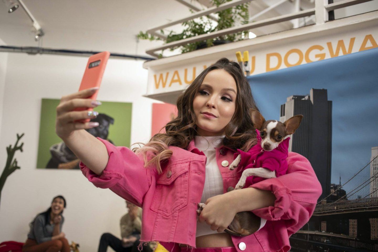 Larissa Manoela vestindo uma jaqueta rosa tira selfie com um cachorro no colo.