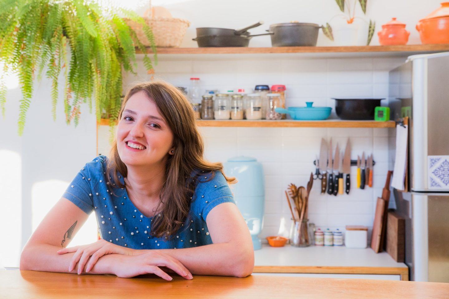 Cristal Muniz à mesa com uma prateleira de objetos e uma samambaia ao fundo.