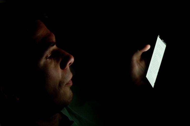 Homem com o rosto parcialmente iluminado mexendo no celular no escuro.