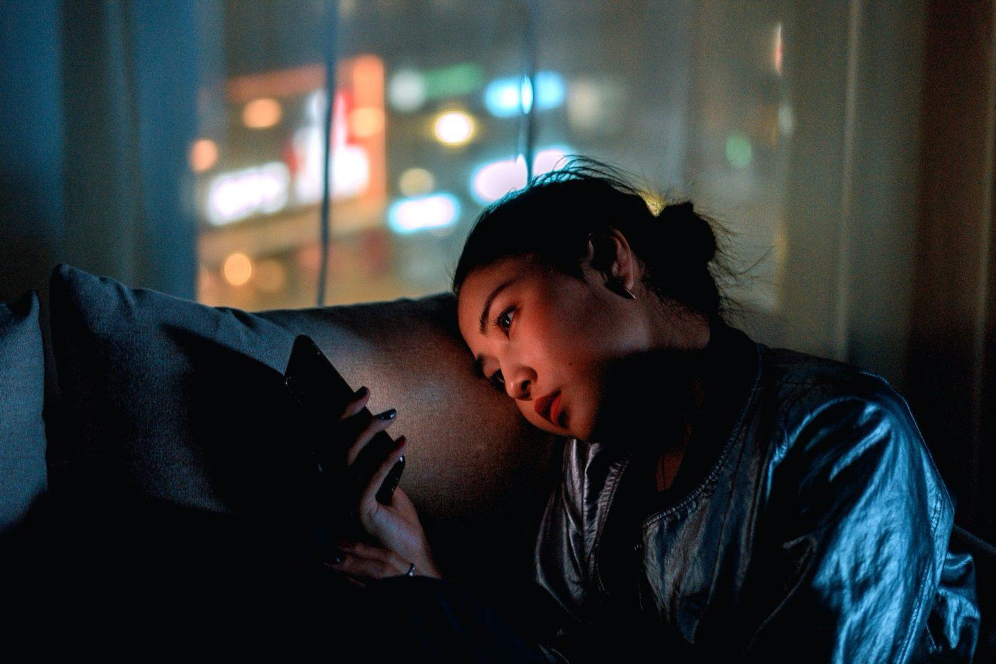 Mulher sentada no sofá olha para celular no escuro, com a tela iluminando seu rosto. Ao fundo, uma janela.