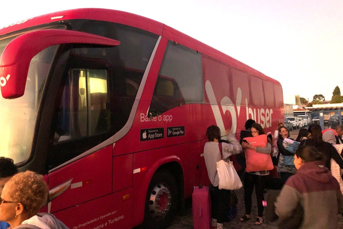 Lateral de um ônibus da Buser com alguns passageiros sonolentos ao lado.