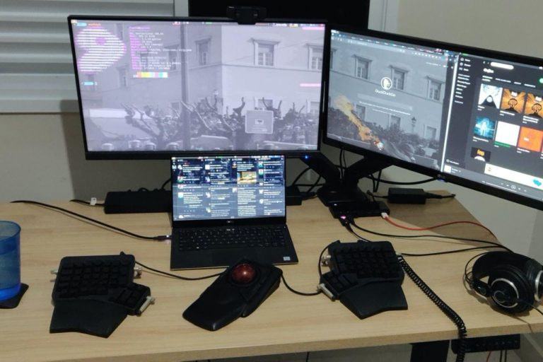 Escritório doméstico do Flávio, com notebook, dois monitores e acessórios ergonômicos sobre a mesa.
