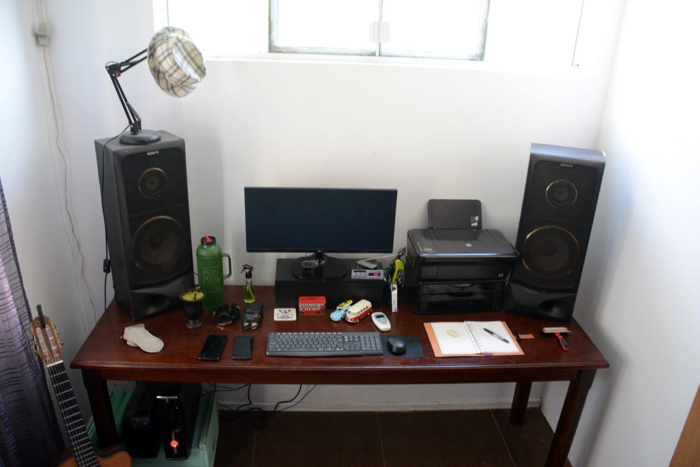 Mesa de trabalho do Rafael Goulart, com computadores, caixas de som grandes, impressora e uma janela com boa iluminação natural acima.