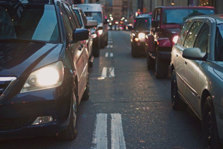 Foto frontal de duas fileiras de carros, faróis ligados, no crepúsculo.