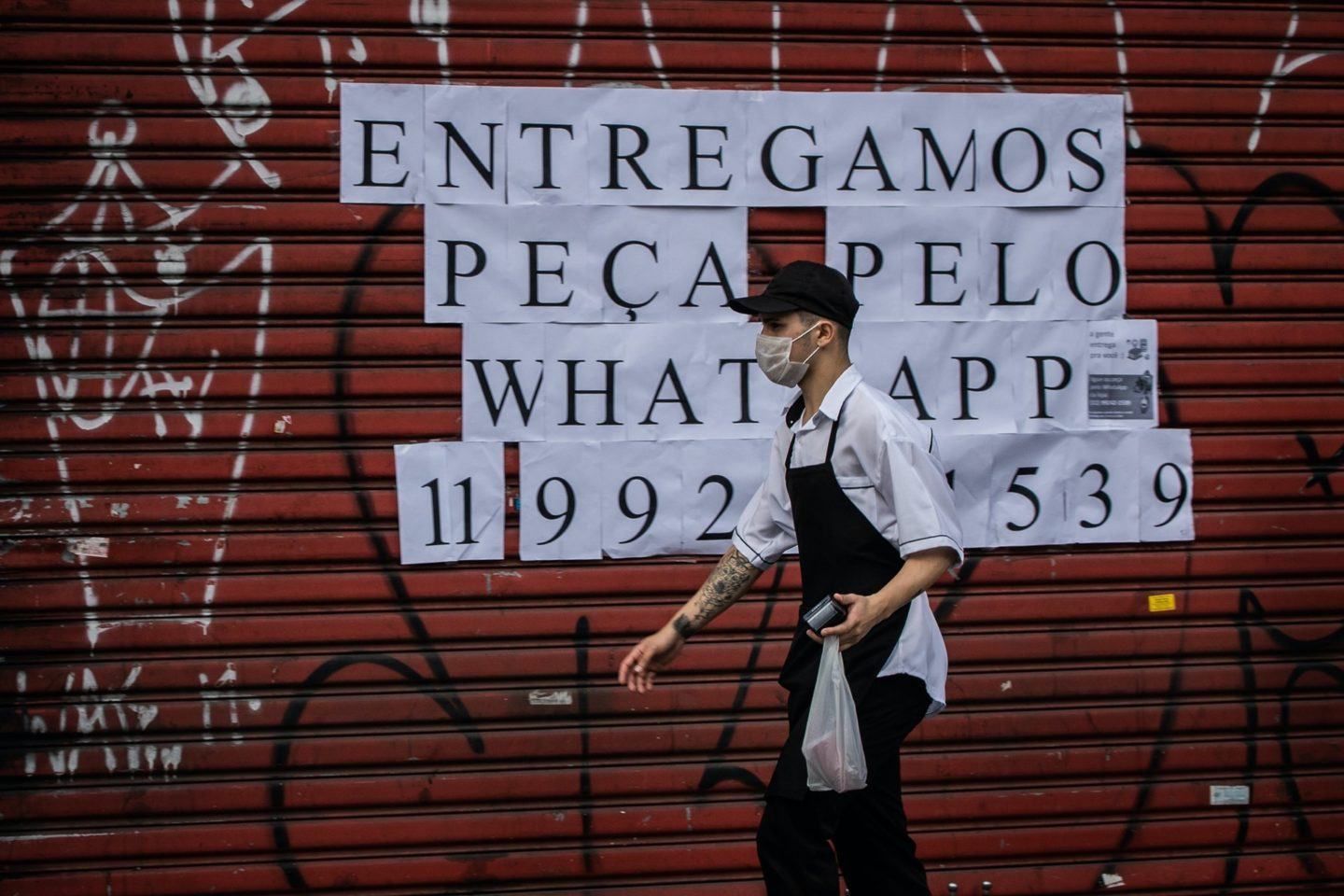 Funcionário de avental carrega uma sacola de refeição; ao fundo, porta de estabelecimento fechada com algumas folhas impressas atrás fazendo propaganda de entregas de um restaurante via WhatsApp.
