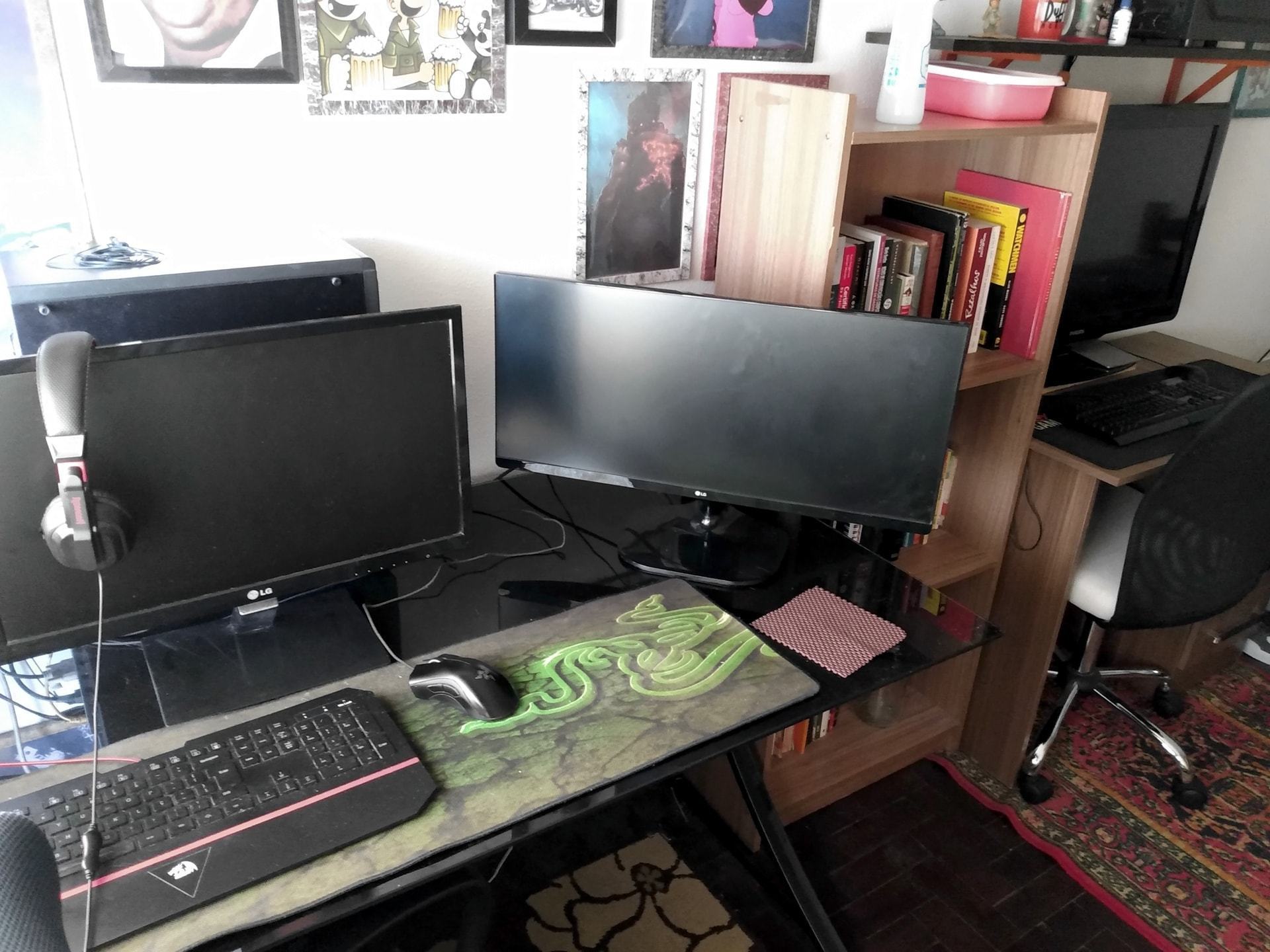 Mesa em primeiro plano com dois monitores, teclado e mouse; ao lado/fundo, estante de livros e a segunda mesa, de jogos, com uma TV enorme.