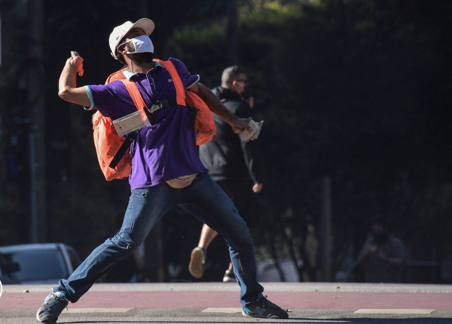 Homem pardo de máscara e com uma mochila de aplicativo de entrega arremessando uma pedra durante manifestação.