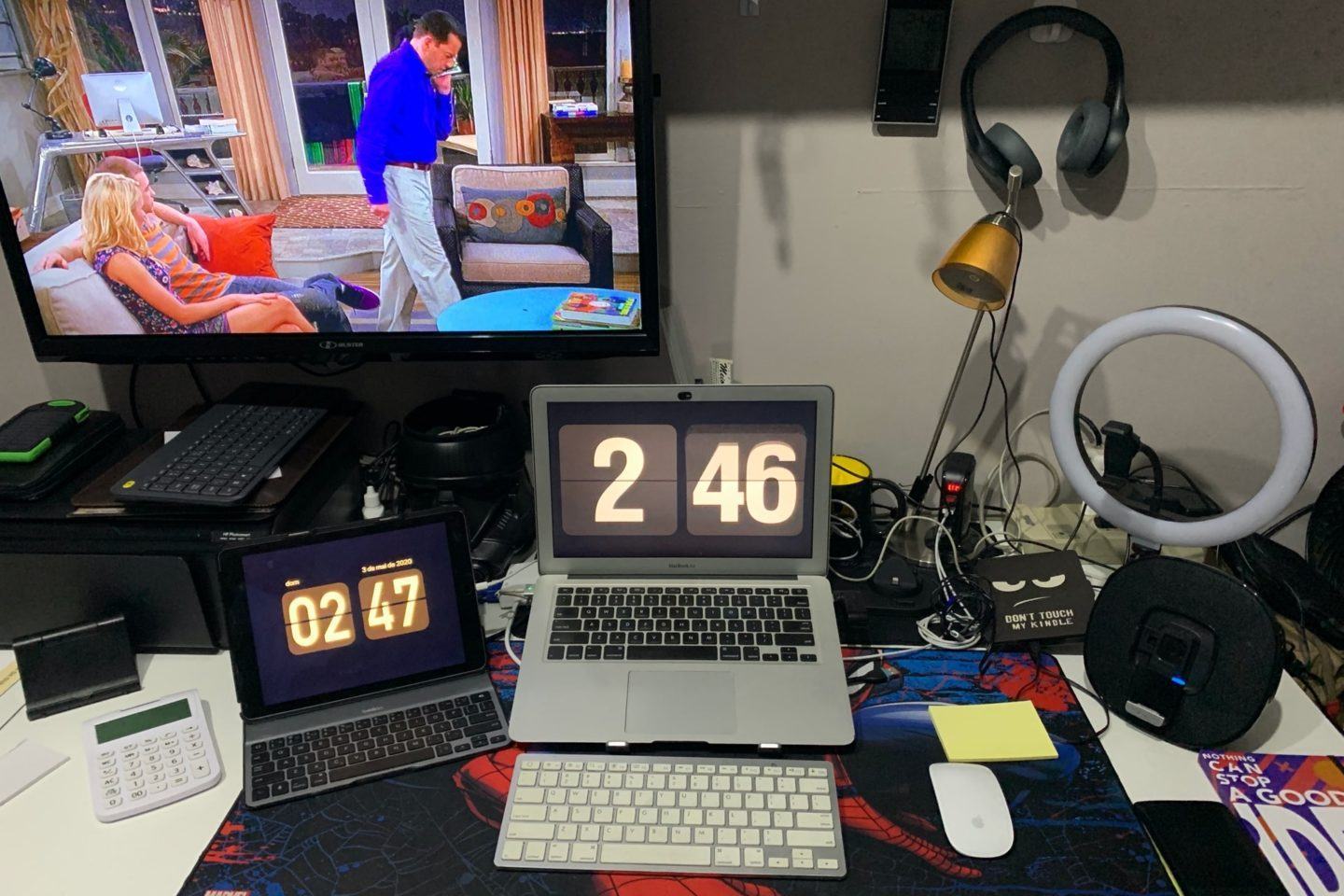 Foto da mesa de trabalho em casa do Uri Barros. Notebook e tablet, ambos com teclados externos; ao fundo, uma TV. Mesa com bastante coisa em cima.