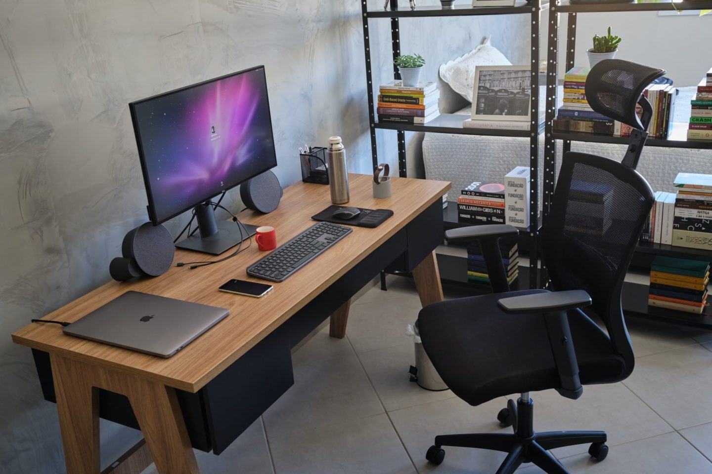 Foto de lado de uma mesa de trabalho, com notebook, monitor grande, teclado, mouse e outros objetos sobre ela. Poltrona à vista e, ao fundo, uma estante vazada.