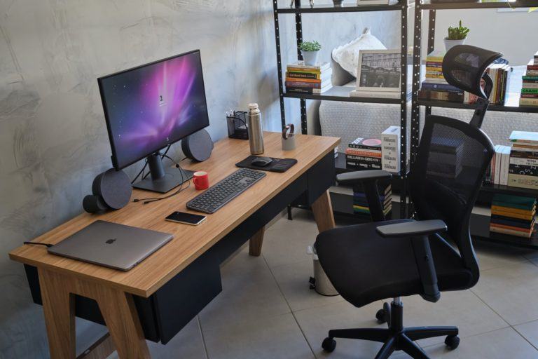 Foto de lado de uma mesa de trabalho, com notebook, monitor grande, teclado, mouse e outros objetos sobre ela. Poltrona à vista e, ao fundo, uma estante vazada.