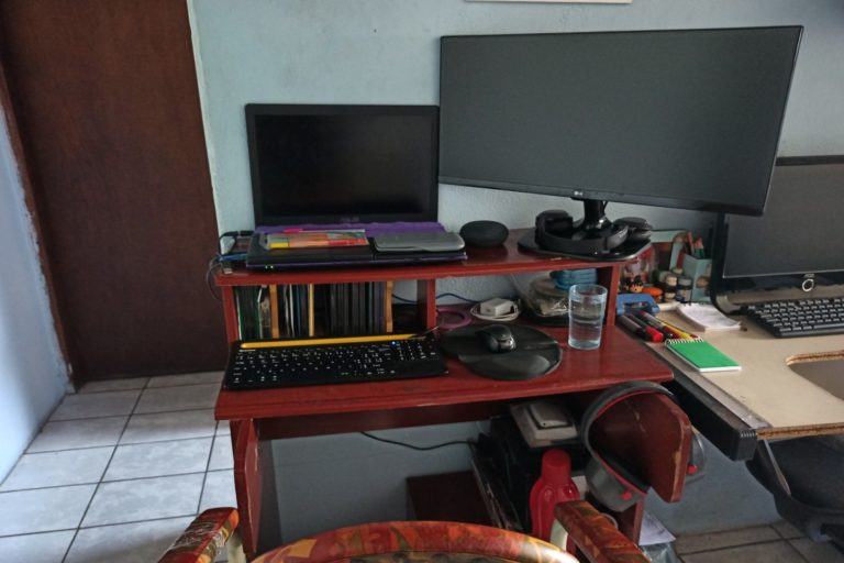 Escritório doméstico do Henrique Bispo, mostrando um rack com notebook, teclado e mouse; um monitor grande e alguns objetos.