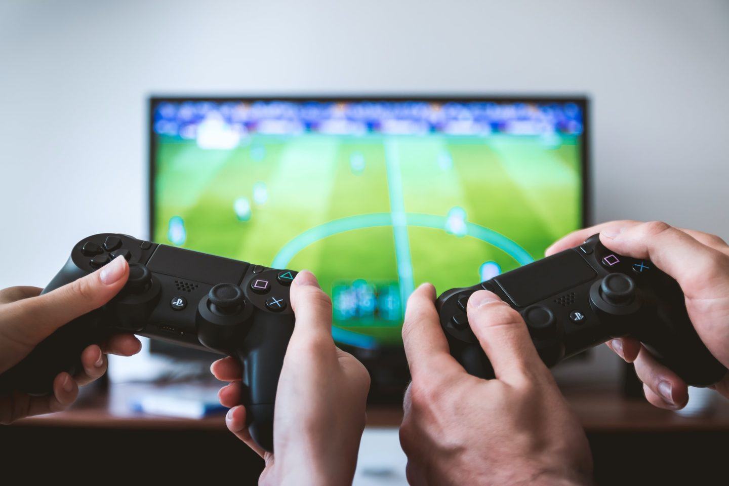 Dois pares de mãos segurando controles de PlayStation e, ao fundo, uma TV exigindo um jogo virtual de futebol.