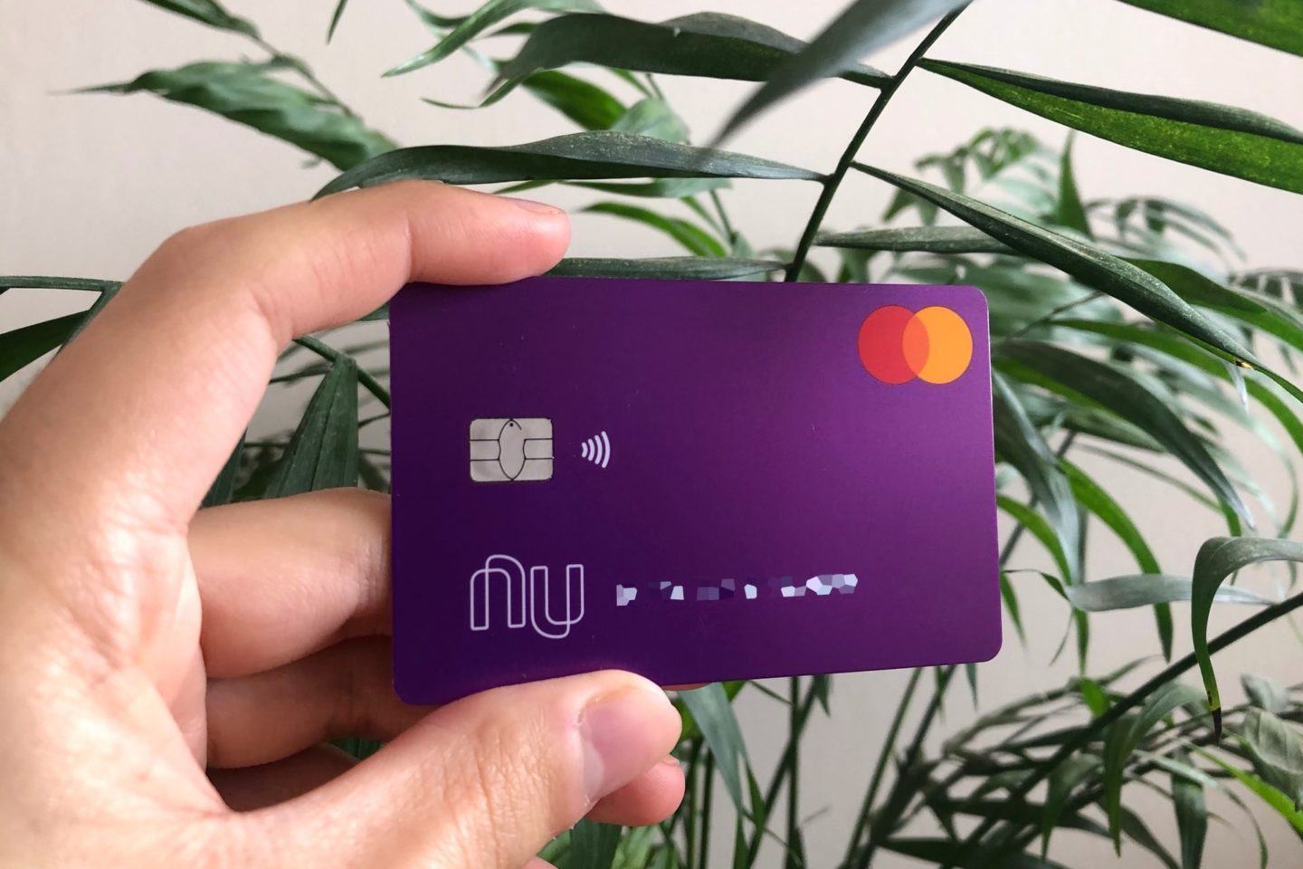 Cartão contactless do Nubank segurado por uma mão contra um fundo de parede branca com uma palmeira.