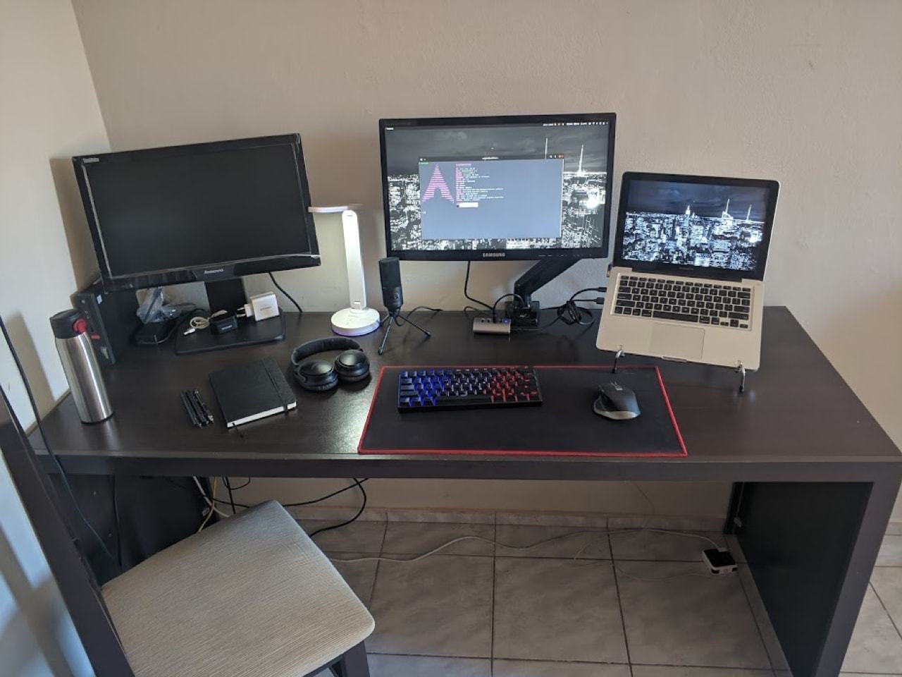 Mesa de trabalho, com uma cadeira de cozinha. Sobre a mesa, um notebook, dois monitores, acessórios espalhados.