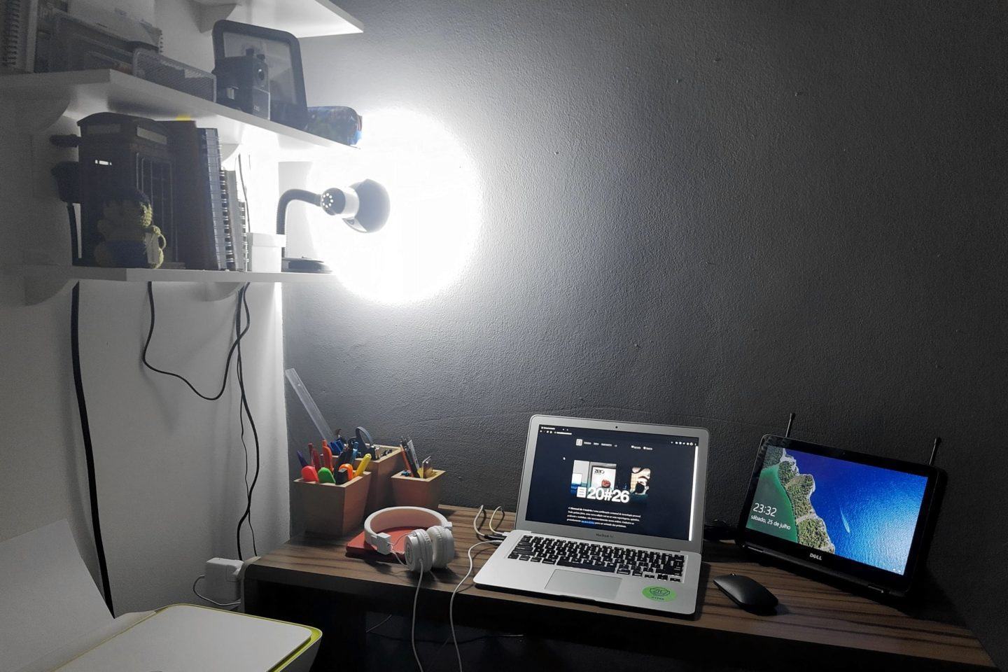 Mesa com dois notebooks, um mouse e um par de fones de ouvido. À esquerda, prateleira com objetos, lâmpada forte e, no canto inferior, um multifuncional branco e amarelo.