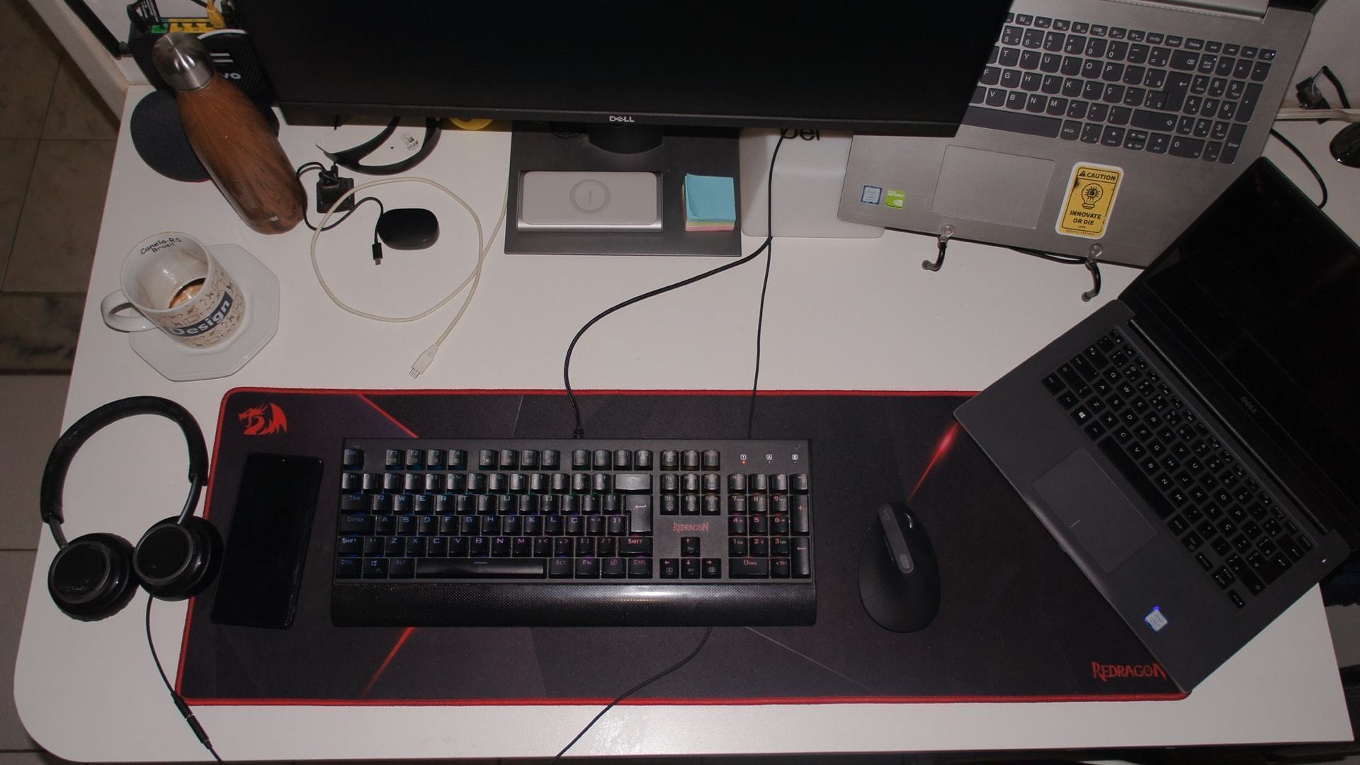 Vista de cima da área de trabalho do Matheus Falcão, com monitor, notebooks, teclado, fones de ouvido e outros objetos.