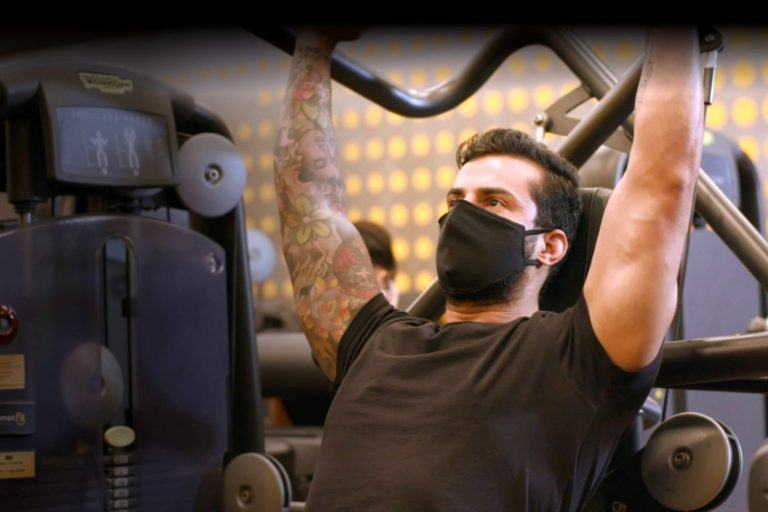 Homem musculoso e com um braço tatuando, usando máscara, levanta peso em um equipamento de musculação.