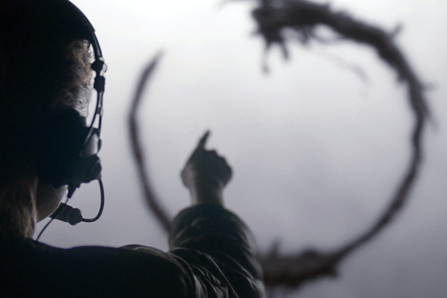Círculo com bordas escuras desenhado em uma parede branca. No lado esquerdo do enquadramento, de frente para o círculo, mulher com headset apontando para o círculo.