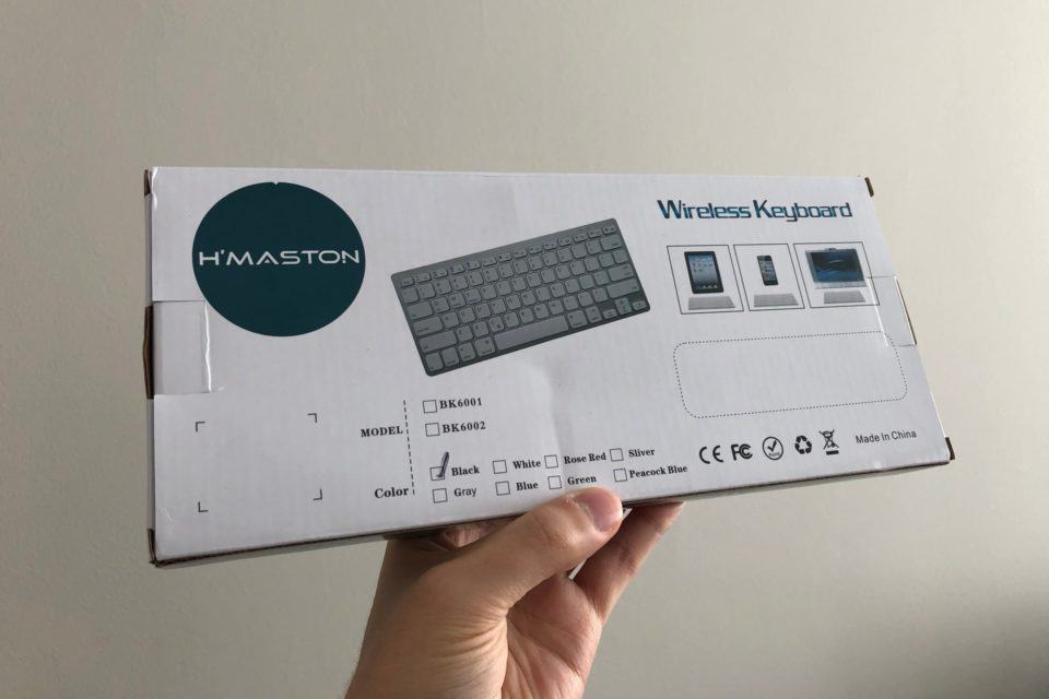 Caixa do teclado Bluetooth H.Maston segurada por uma mão contra uma parede branca.
