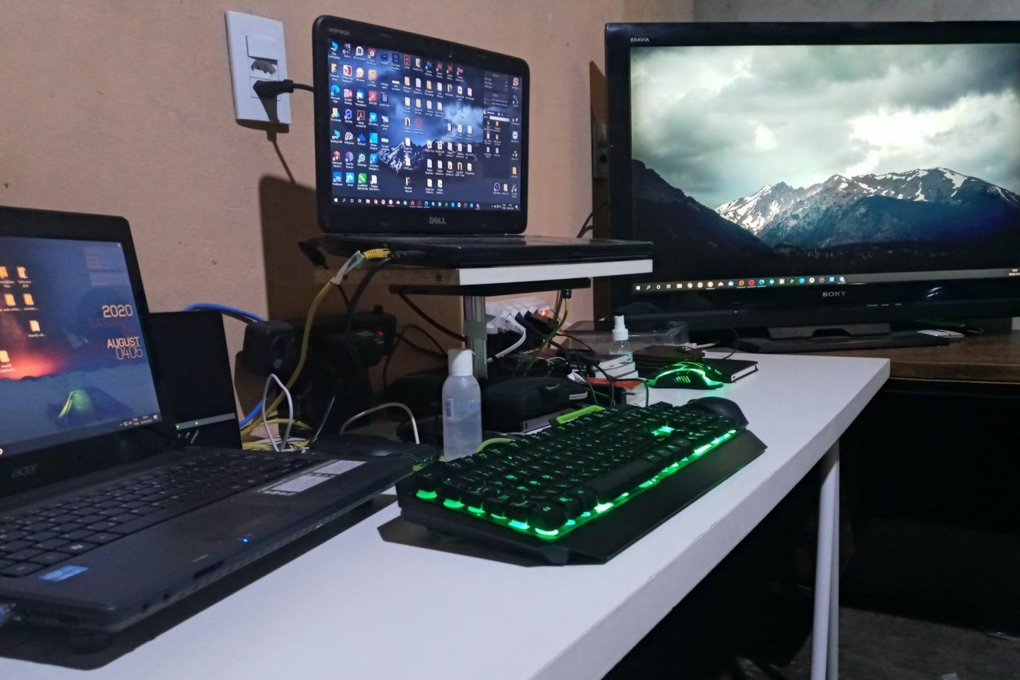 Foto de uma mesa, de lado, com dois notebooks, um deles elevado, teclado e mouse iluminados na cor verde e, ao fundo, uma TV grande.