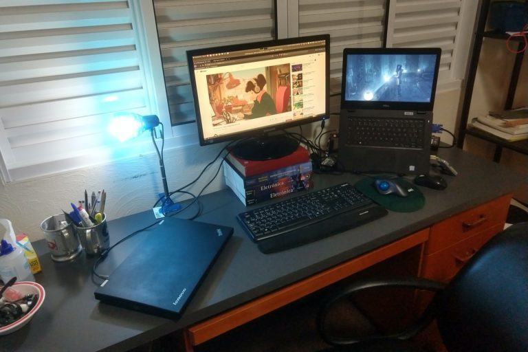 Foto de lado da mesa do Caio, com monitor, dois notebooks (um fechado e outro aberto) e acessórios. Ao fundo, uma janela fechada e uma lâmpada forte sobre a mesa.