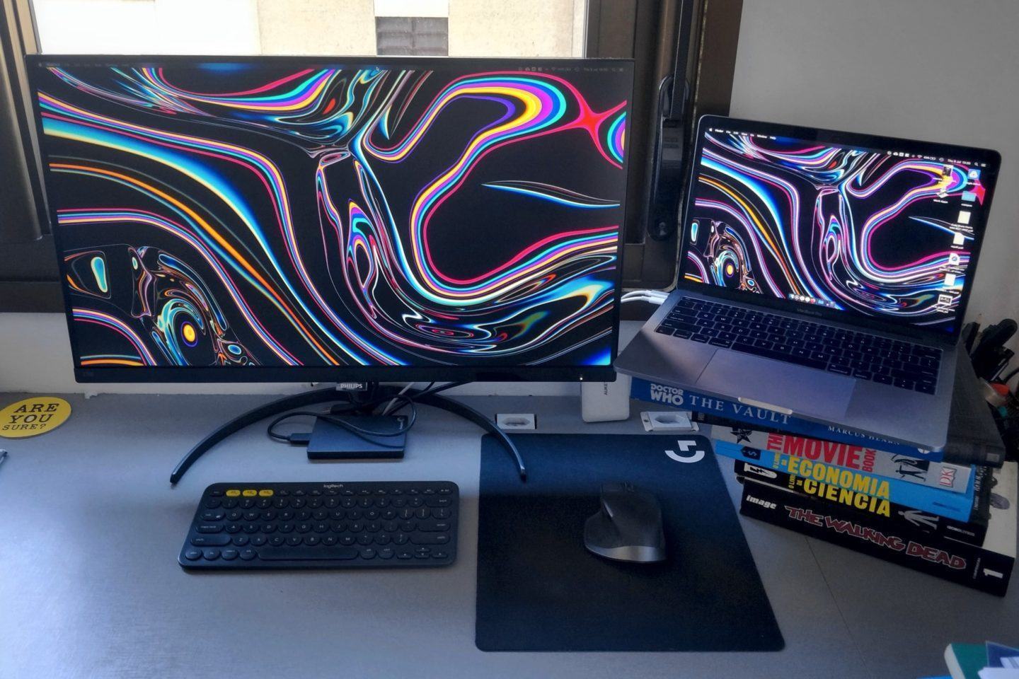 Mesa de trabalho com um notebook à direita sobre uma pilha de livros, um monitor grande ao lado, teclado, mouse e um grande mousepad.