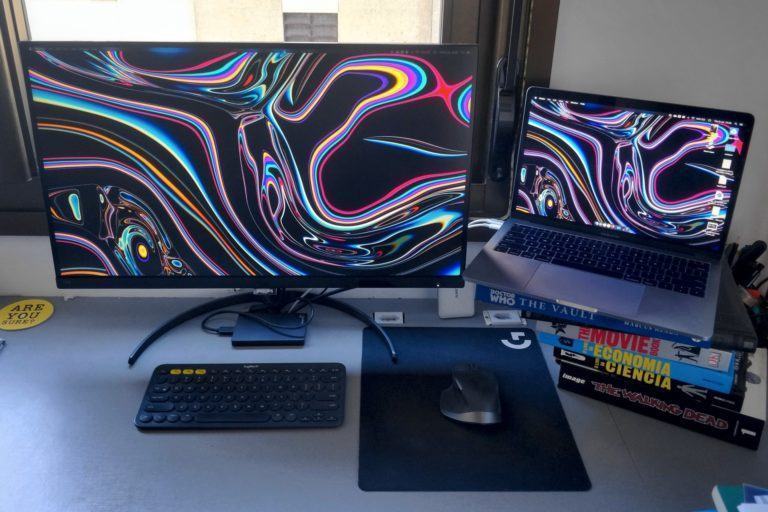 Mesa de trabalho com um notebook à direita sobre uma pilha de livros, um monitor grande ao lado, teclado, mouse e um grande mousepad.