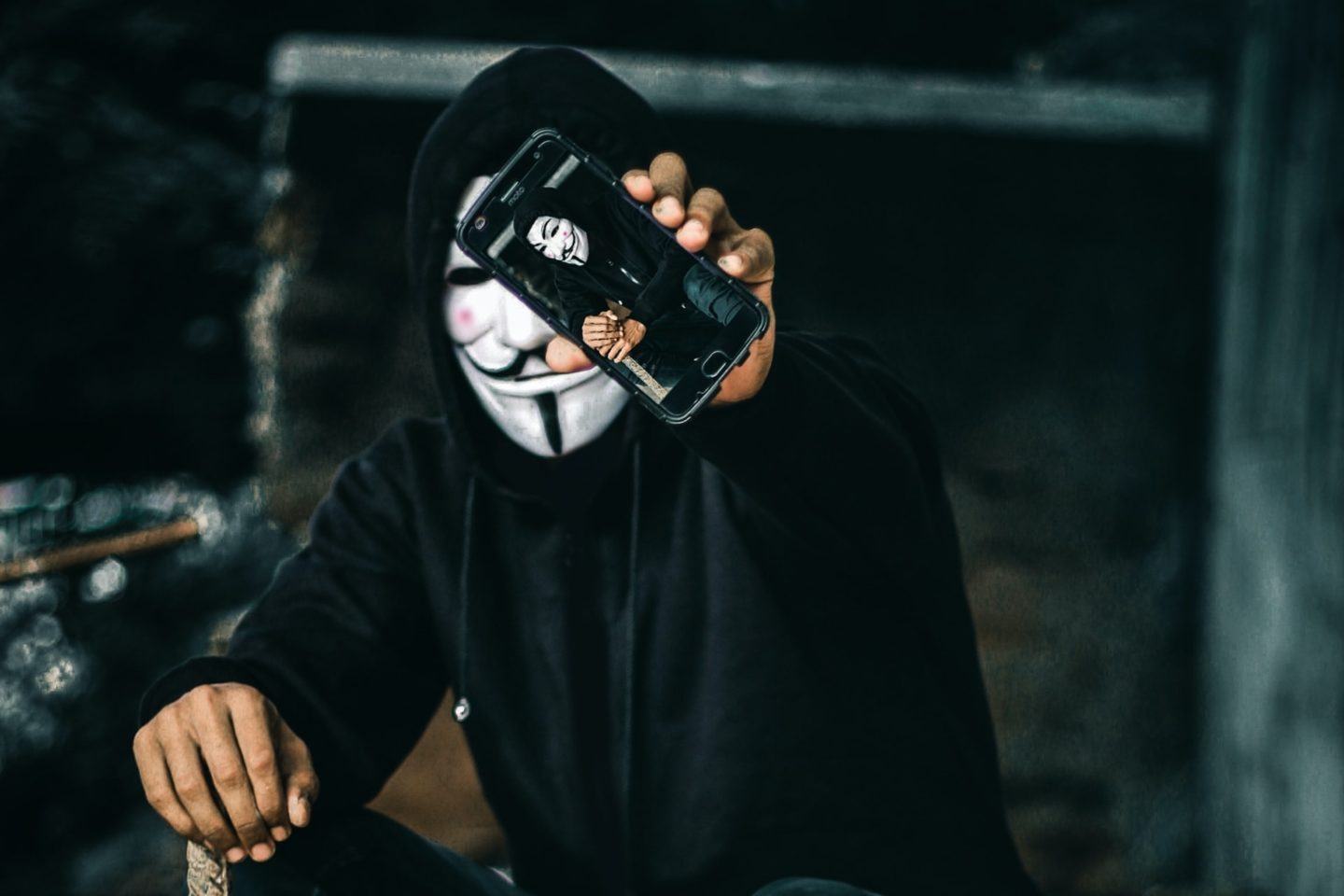Homem vestido de preto com a máscara do Guy Fawkes segurando um celular mostrando a tela que o enquadra na câmera.