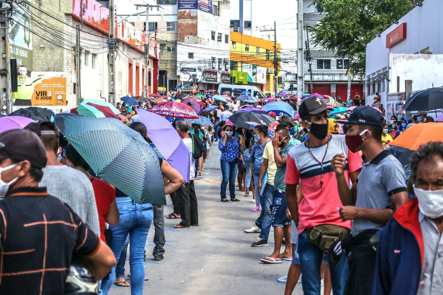 Pessoas de máscara e segurando guarda-chuvas em uma grande fila no meio da rua.