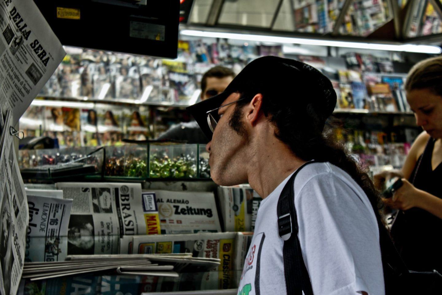 Homem de boné, barba e óculos, de lado, olha jornais pendurados na parede de uma banca.