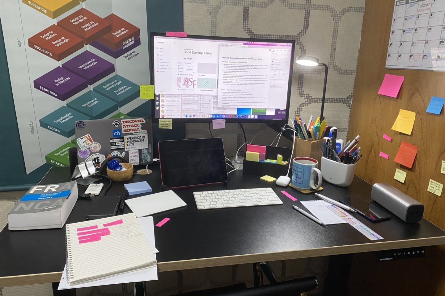 Foto de uma mesa, com monitor, teclado, mouse e notebook fechado, de pé, uma série de papéis e outros pequenos objetos. Na parede à direita, de madeira, vários post-its.