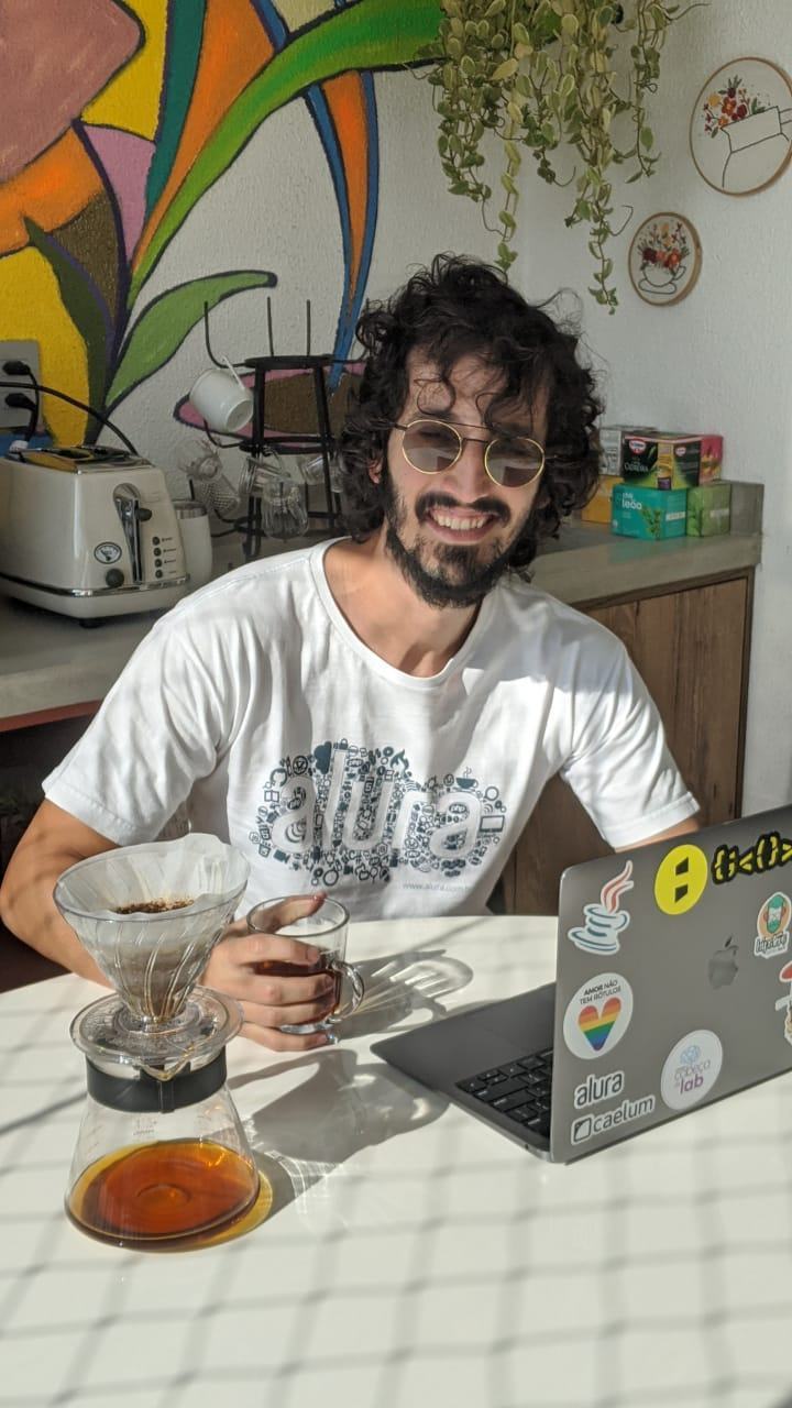Homem sorridente, de óculos, cabelos encaracolados e barba, sentado à mesa com uma xícara de café na mão e um notebook à sua frente.