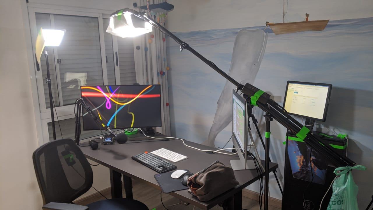 Mesa com uma luminária apoiada em tripé sobre ela, um monitor grande e um iMac (um em cada ponta) e acessórios espalhados.