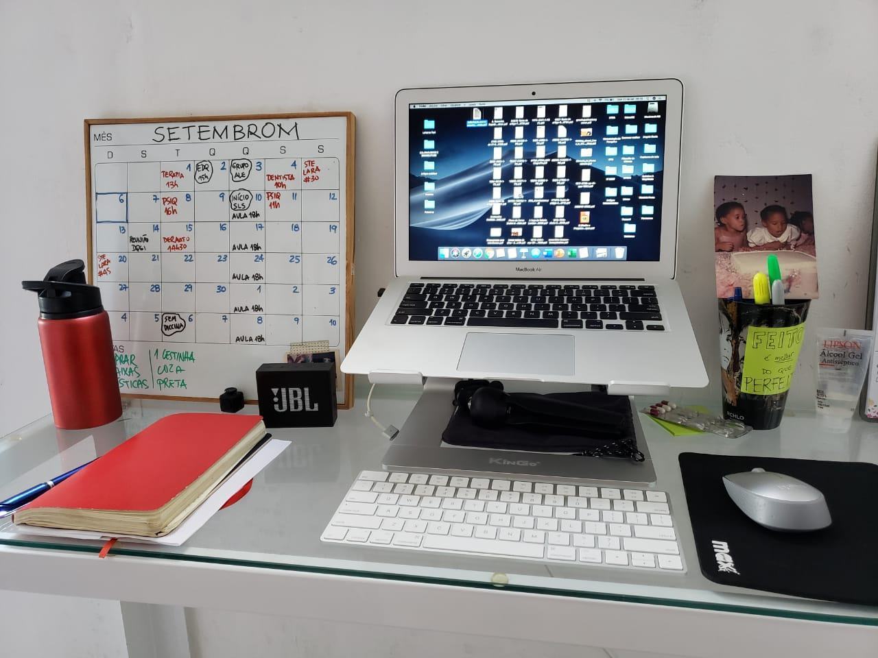 Mesa clara, com um MacBook Air em um suporte, teclado e mouse, quadro branco com anotações e outros objetos espalhados.