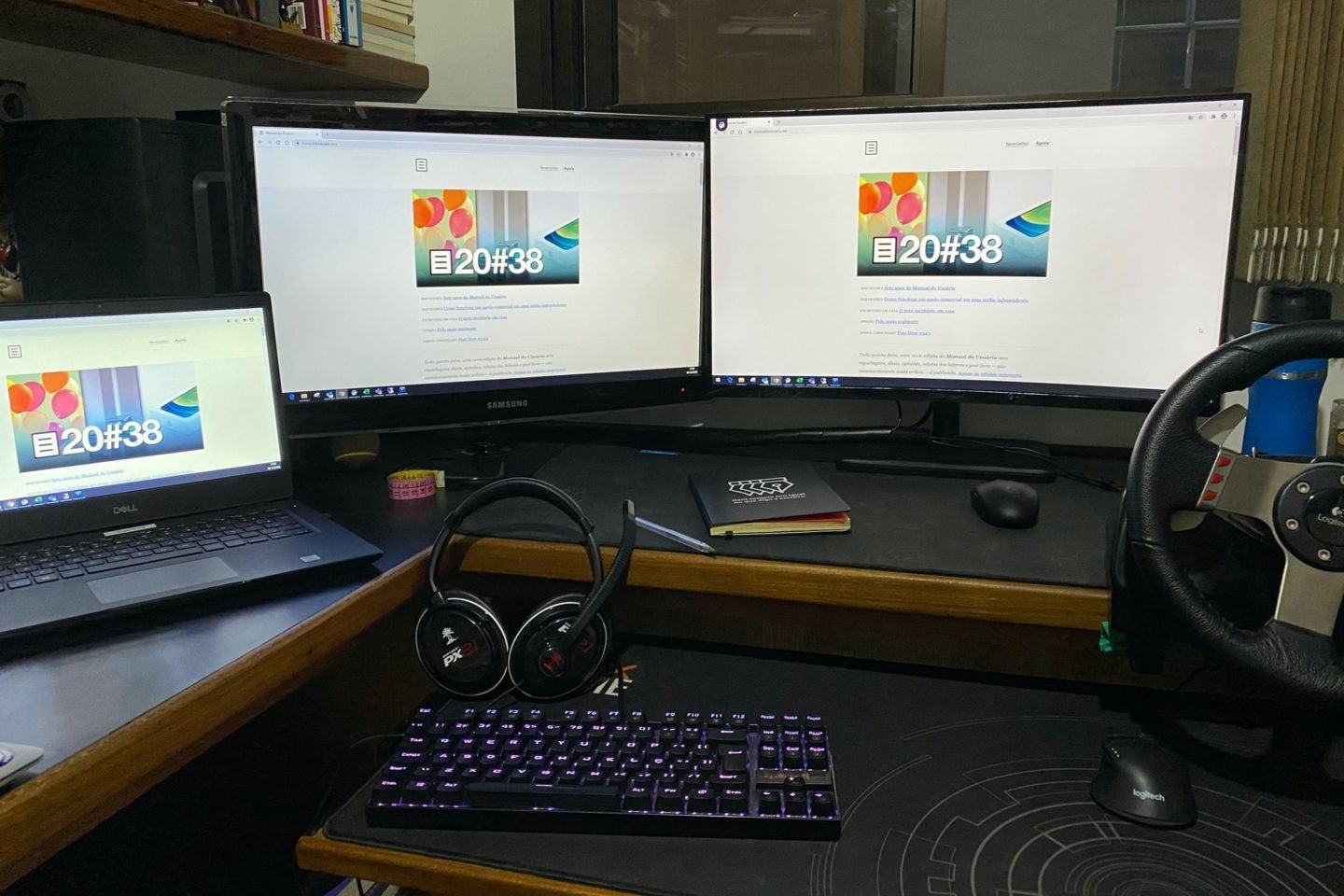 Mesa com dois monitores e um notebook aberto à esquerda, todos com a capa do Manual do Usuário em exibição.