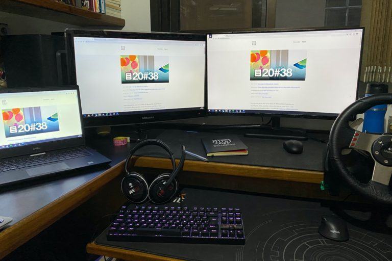 Mesa com dois monitores e um notebook aberto à esquerda, todos com a capa do Manual do Usuário em exibição.