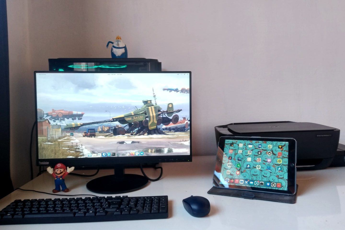 Monitor grande com um teclado, mouse e boneco do Mario à frente; atrás dela, um computador com personagem da Hora de Aventura em cima. À direita, iPad com a tela ligada e uma impressora preta ao fundo.