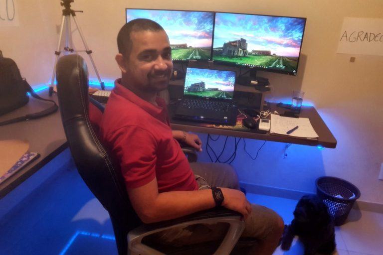 Homem de barba e cabeça raspada sentado em uma cadeira gamer, de lado, olhando para a câmera. Ao fundo, mesa com dois monitores grandes e um notebook entre eles, e várias coisas espalhadas sobre.