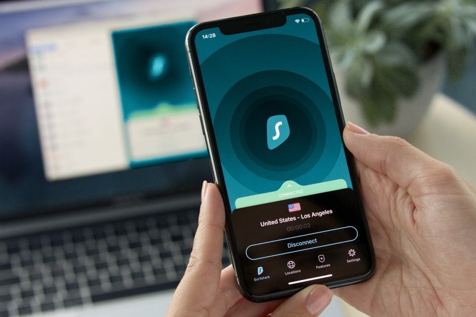 Celular com o SurfShark aberto segurando por duas mãos; ao fundo, um notebook desfocado, também com o SurfShark aberto.