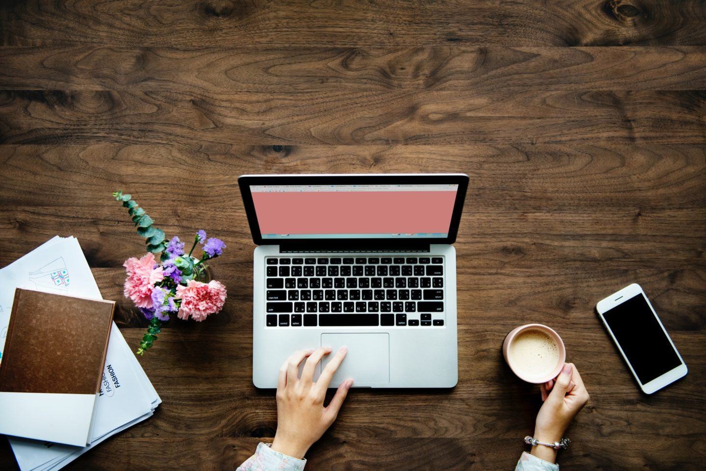 Foto de cima de uma mesa, com mãos mexendo em um computador e segurando uma xícara de café. Nas laterais, um celular e alguns livros.