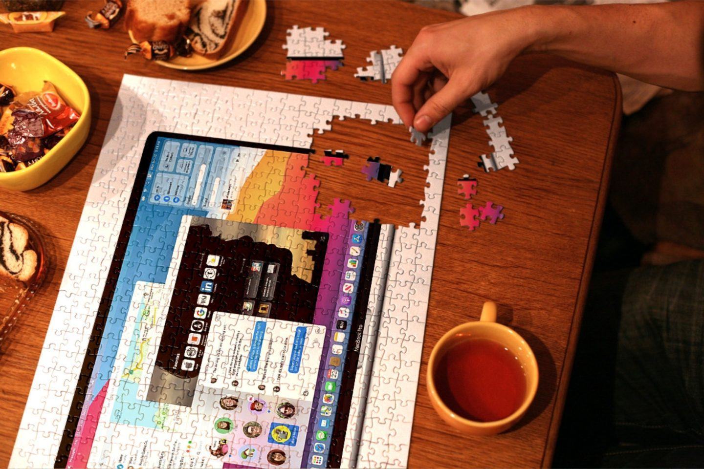 Quebra cabeça com algumas peças faltando de um notebook da Apple com o macOS Big Sur. Ao redor, caneca com café, uma mão, algumas peças e chocolates.