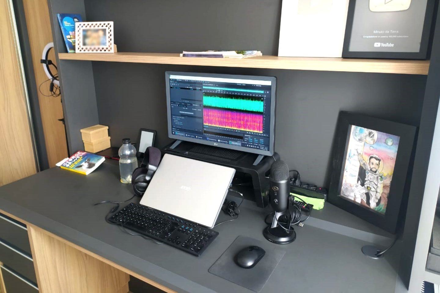 Mesa escura com notebook fechado, monitor, teclado, mouse e decorações.