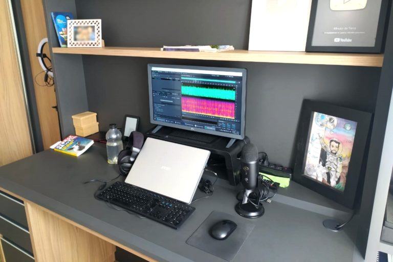 Mesa escura com notebook fechado, monitor, teclado, mouse e decorações.