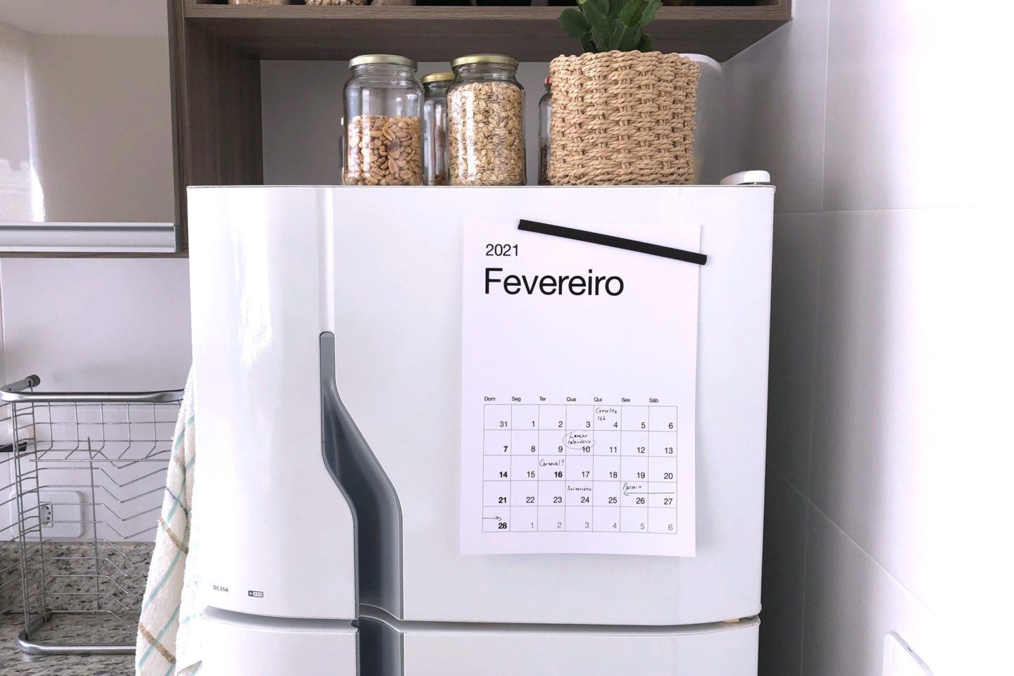Foto de uma geladeira branca, com alguns potes de oleaginosas e uma planta em cima. Na porta do congelador, um folha com o calendário de fevereiro de 2021.